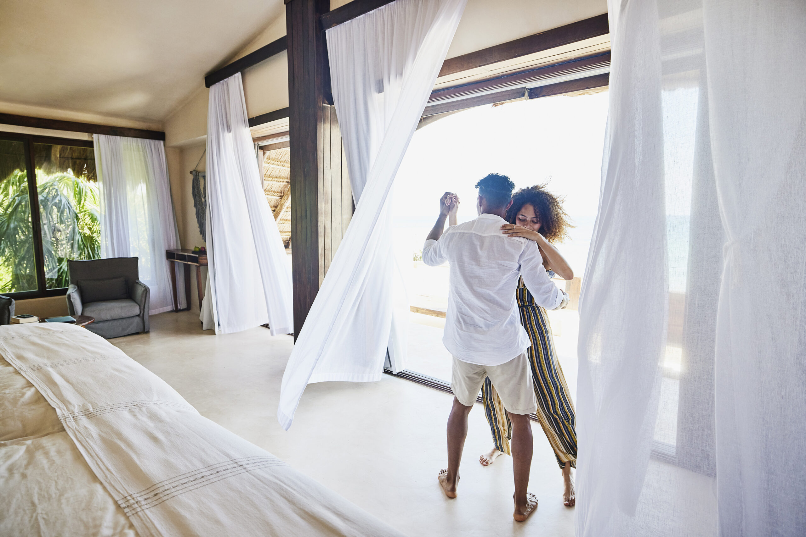 Foto panorâmica de um casal dançando em uma suíte de luxo de um resort tropical.