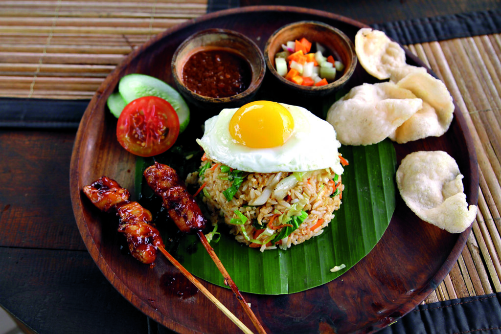 Arroz frito picante con gambas, verduras, pollo frito, galletas de pescado, pepinillos y satay de pollo, coronado con huevo frito y sambal de chile, anchoas y vieira seca