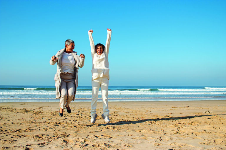 Two excited female friends having fun at seashore on sunny autumn day. Senior ladies laughing and jumping with raised arms while having great vacation time at seaside. Vacation, joy, friends concept